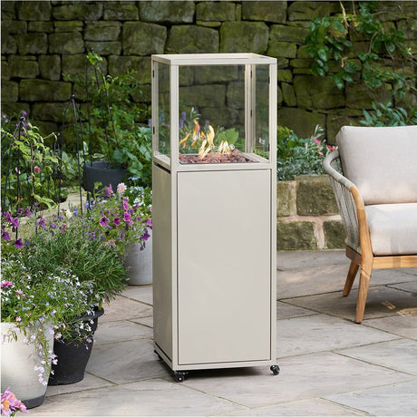 Cosi Dome Sandy Beige Gas Lantern, 116cm tall with aluminium frame and glass panels, lit flame with lava rocks, on a patio beside potted plants and a stone wall.