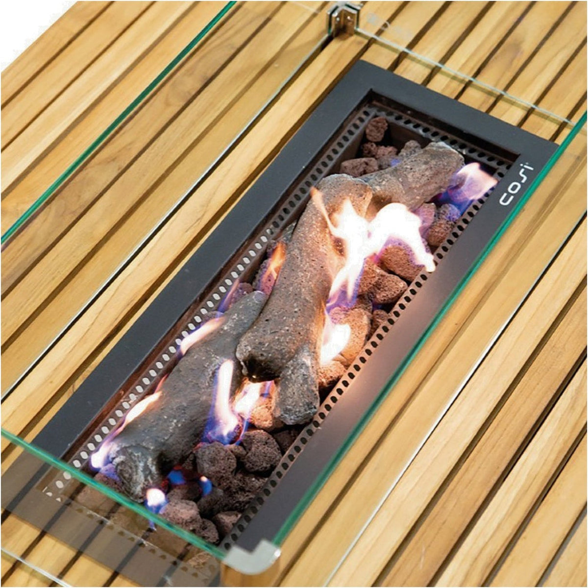 Cosi Gas Fire Pit Table close-up showing lit lava rocks and ceramic logs within a black burner insert, framed by glass wind guard and teak slatted top.