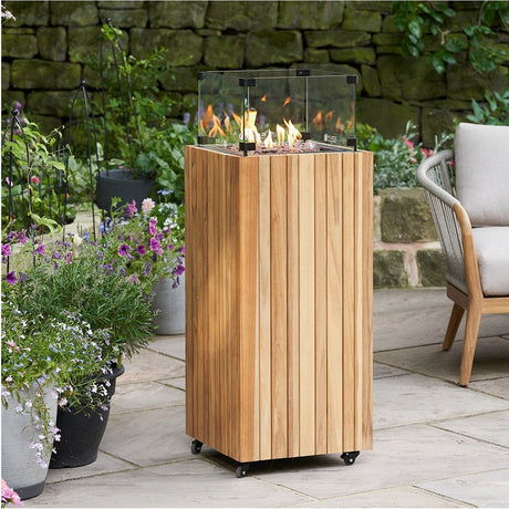 Cosi Teak Gas Lantern – 105cm, tall wooden-clad fire lantern with glass surround and flame visible, on castors in a garden patio setting.