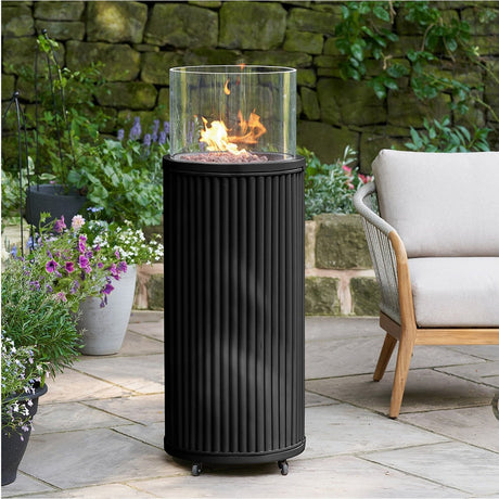 Cosi Tube Black Gas Lantern, 106cm, with fluted aluminium cylindrical base and glass cylinder top with lit flame, on wheels, shown on a garden patio beside a chair and plants.