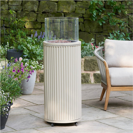 Cosi Tube Sandy Beige Gas Lantern, 106cm tall with ribbed aluminium base and clear glass tube, shown on a patio beside plants and outdoor seating.