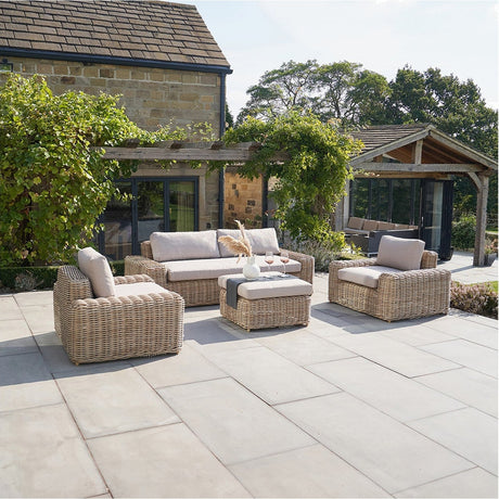 Como Natural Antique outdoor seating set with rattan-effect sofa, two armchairs, and a footstool table on a patio, shown against a stone house with pergola.