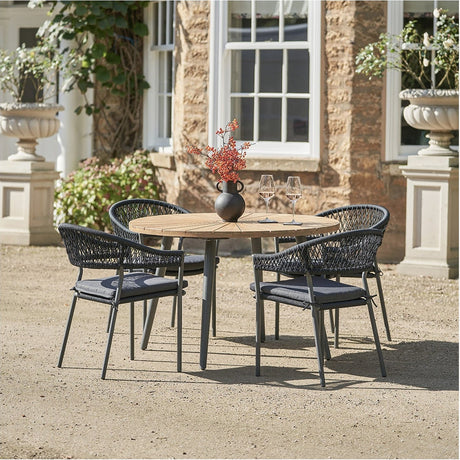 Bazel Outdoor 4 Seater Dining Set with round wood-top table and black woven aluminium chairs, set outdoors on gravel against a stone house facade.