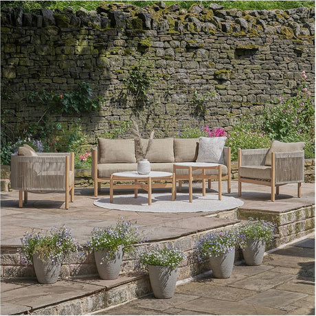 Marche Outdoor Seating Set with a wood-framed sofa, two armchairs, and round nested tables in beige woven and cushioned fabric, set on a stone patio against a rustic stone wall.