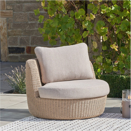 Kampala Outdoor Lounge Chair with round woven beige base, beige cushion seat and backrest, set outdoors on a patio with a rug and greenery backdrop.