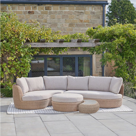 Kampala Outdoor Corner Set curved rattan-effect sofa with beige cushions, round ottoman and side table, shown on a patio against a stone wall and greenery.