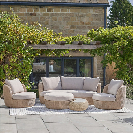Kampala Outdoor Lounge Set with woven rattan-effect sofa, two swivel chairs, oval coffee table and stool, beige cushions, set on a patio against a stone house and greenery.