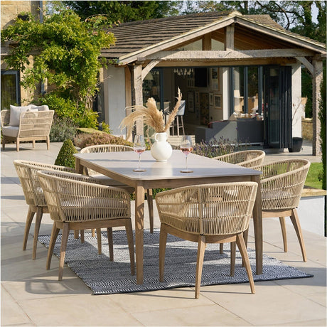 Moeven Outdoor 6 Seater Dining Set with rectangular composite-top table and woven wicker armchairs on wooden legs, shown on a patio beside a house.