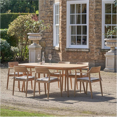 Salita Outdoor 6 Seater Dining Set in natural wood, oval table with six armchairs and beige cushions, set on gravel outside a stone house.