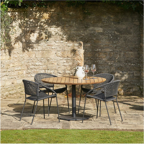 Dakota Black Outdoor 4 Seater Dining Set with round teak-top table and woven black chairs on a stone patio against a rustic wall.