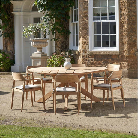Reuleaux Outdoor 6 Seater Dining Set in natural teak, round table with cushioned wooden armchairs, set outdoors on gravel before a stone house.