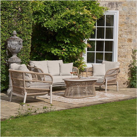 Martic Outdoor Seating Set in beige woven aluminium, with a three-seat sofa, two armchairs, and round glass-top coffee table, shown on a patio against a stone house and greenery.