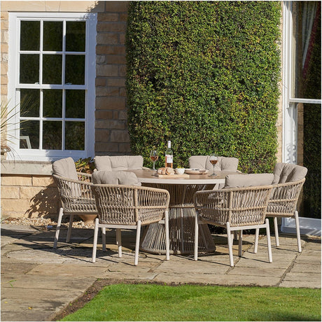 Martic Outdoor 6 Seater Dining Set with round glass-top table and woven beige rattan armchairs with cushions, set on a garden patio against a green hedge wall.