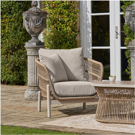 Martic Outdoor Chair with woven latte polypropylene frame and beige cushions, set on a patio beside a matching round coffee table in a garden setting.