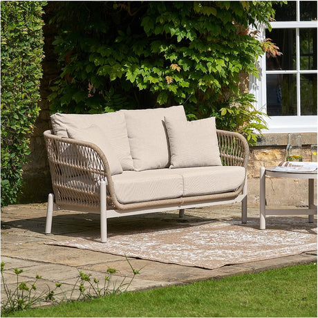 Martic Outdoor 2 Seater Sofa in beige, with woven rope frame, aluminium legs, and cushioned seat and back pillows, shown on a garden patio beside a matching side table.