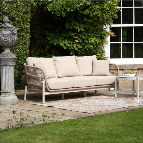 Martic Outdoor 3 Seater Sofa with rounded woven latte frame, aluminium legs, and cream cushions, shown on a patio beside a garden wall and window.