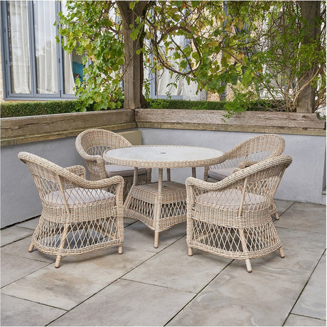 Pattaya Outdoor 4 Seater Dining Set featuring a round 85cm table with four woven aluminium armchairs in sand, set on a patio surrounded by greenery.