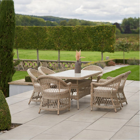 Pattaya Outdoor 6 Seater Dining Set in sand-beige synthetic rattan weave with aluminium frame, glass-top table and cushioned chairs, on a patio overlooking a lawn and hedges.