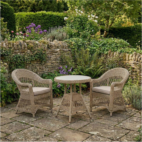 Pattaya Outdoor Bistro Set with round glass-top table and two woven armchairs in sand-beige synthetic rattan, set on a garden patio with stone walls and greenery.