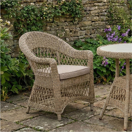Pattaya outdoor dining chair with curved woven rattan-effect back and arms in sand, aluminium frame, and beige cushion, shown on a patio beside a matching round table.