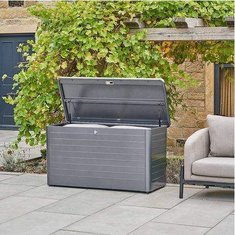 Rhode Island galvanised steel cushion box in anthracite, lid open revealing cushions inside, shown on a patio beside a sofa and greenery.