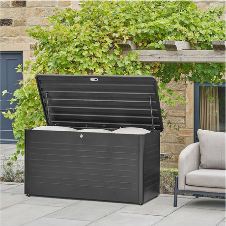 Rhode Island large galvanised steel cushion box in black, lid open with cushions inside, ridged panel design, shown outdoors on a patio near a garden wall.