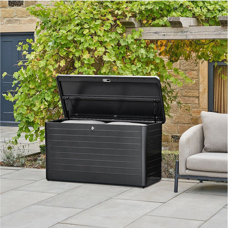Rhode Island Small Galvanised Steel Cushion Box in black, open on a patio with cushions inside, set against a stone wall with climbing greenery.