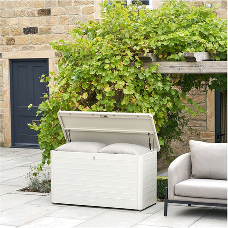 Rhode Island Small galvanised steel garden cushion box in pebble, open lid revealing grey cushions, shown on a patio beside a sofa and greenery.