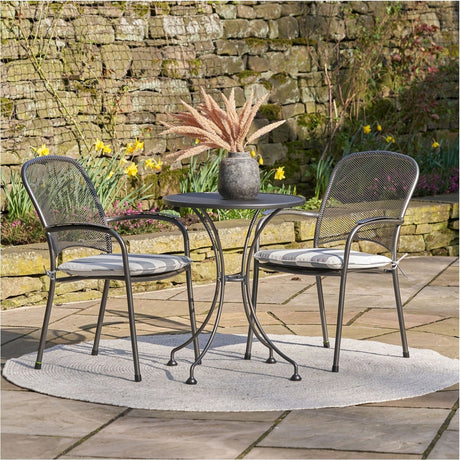 Carlo Bistro Set with Cushions in grey mesh steel, round table and two chairs with striped seat pads, shown on a patio beside a stone wall and garden border.