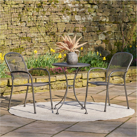 Carlo Bistro Set 89cm garden furniture in iron grey steel, featuring a round mesh table and two matching mesh chairs on a patio beside a stone wall and flowerbed.