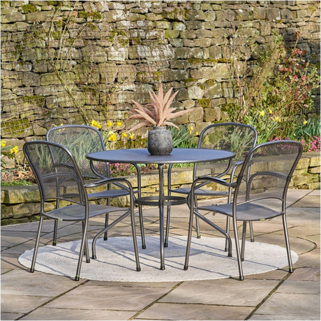Carlo 4 Seater Dining Set in iron grey mesh steel, round table with four stacking armchairs on a patio beside a stone wall and flowerbed.