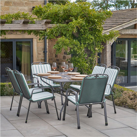 Savoy Rectangular 6 Seater Dining Set on a patio, with a dark metal frame, glass-effect table, and stackable chairs featuring green wicker backs and sage green striped cushions.