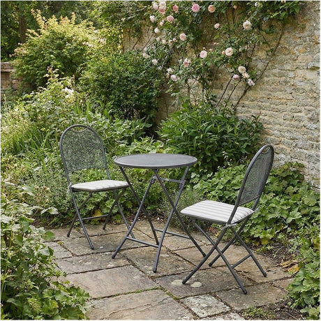 Cafe Latte Balcony folding round set in grey metal with mesh chair backs and grey striped cushions, shown on a garden patio surrounded by greenery.