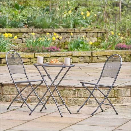 Café Latte Balcony Folding Set - 60cm round table and two folding mesh chairs in grey steel, shown on a patio with a stone wall and garden flowers behind.