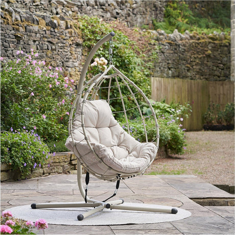 Lyon Putty outdoor hanging egg chair with teardrop rattan-style frame, cushioned seat, and matching stand, shown on a garden patio with a stone wall backdrop.