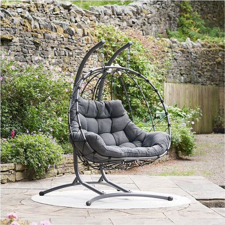 Lyon Anthracite Outdoor Double Hanging Chair with grey cushioned seat, steel egg-shaped frame and stand, set on a patio beside a stone garden wall.
