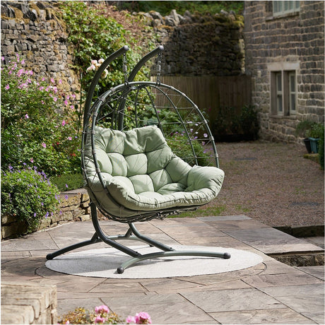 Lyon Moss Green double hanging egg chair with woven grey frame and cushioned green padded seat, set on a stone patio beside a garden wall.