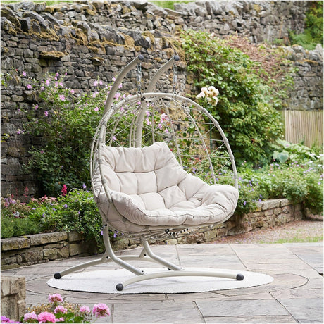 Lyon Putty Outdoor Double Hanging Chair with stand, woven egg-shaped design and cushioned seat, set on a garden patio beside a stone wall.