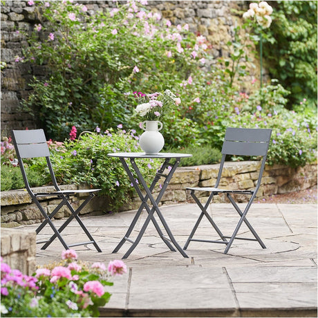 Rennes Anthracite Outdoor Bistro Set with round folding table and two folding chairs in steel, shown on a patio surrounded by garden greenery and flowers.