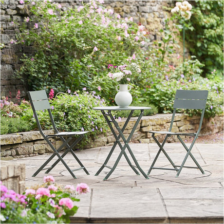 Rennes Moss Green Outdoor Bistro Set, 71cm round metal table with two folding chairs, shown on a garden patio surrounded by flowering shrubs.
