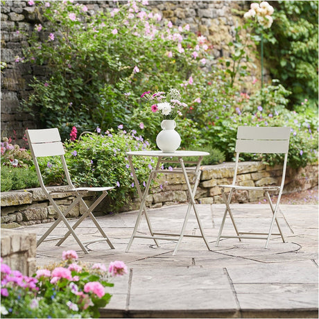 Rennes Putty Outdoor Bistro Set with round folding table and two folding chairs in cream steel, shown on a garden patio with a flower-filled backdrop.