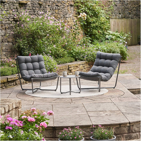 Clermont Anthracite Outdoor Lounging Set with two low-slung metal-framed chairs and grey tufted cushions around a round side table, shown on a stone garden patio.