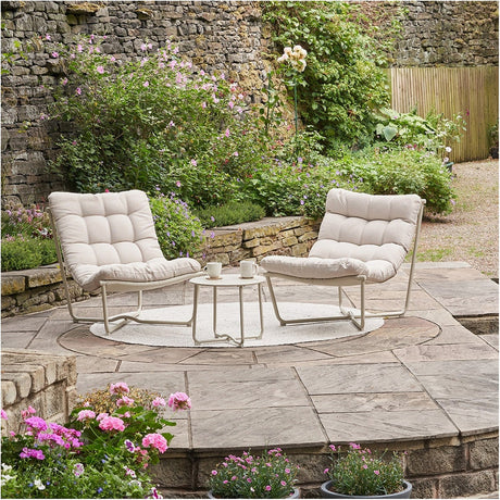 Clermont Putty Outdoor Lounging Set with two low-slung steel frame chairs and tufted putty cushions, plus a round 40cm side table, set on a garden patio.