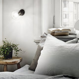 A cozy bedroom corner features white bedding and gray pillows, accompanied by a wooden side table holding a small potted plant. An open book is placed on the windowsill near the modern Prismis Wall Light - Black, which has a black enamel finish and a clear bulb that provides soft illumination.