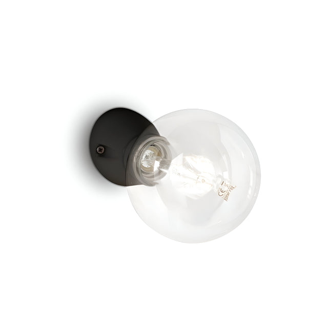 The Prismis Wall Light - Black presents a contemporary design with its black enamel finish and clear round glass bulb, elegantly mounted on a white wall. The transparent bulb reveals the filament within, enhancing the fixture's minimalist and sleek aesthetic.