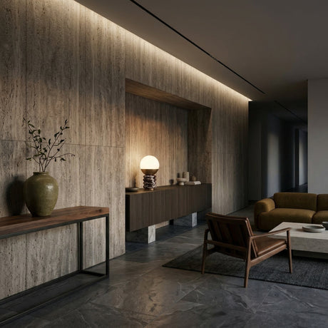 A modern living room with stone walls and floors featuring a wooden console table with a green vase, a brown armchair, a green sofa on a gray rug, and the Velar 1 Light Sculpted Marble Table Lamp in Brass & Purple Red on the sideboard.