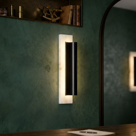 The Velorin LED Small Compact Alabaster Wall Light in soft white and black is mounted on a green textured wall near a mirror and wooden shelf, adding a modern accent with its black cylindrical design and glowing rectangular backplate.