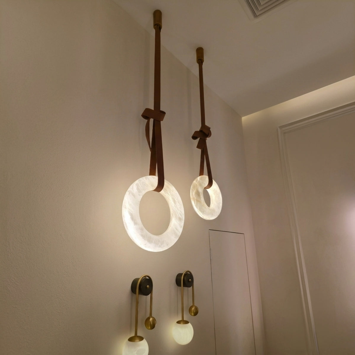 Cintura LED alabaster ring chandelier in warm white, two sizes shown suspended from brown leather straps with copper fittings above a hallway sconce.