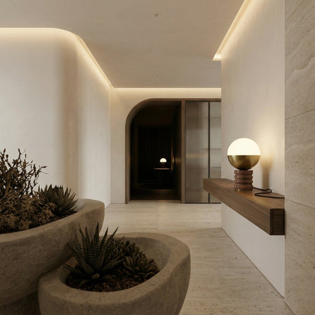 A modern, minimalist hallway with smooth beige walls, a floating wooden shelf holding the Velar 1 Light Sculpted Marble Table Lamp in Brass & Dark Terracotta Red, large stone planters with succulents, and soft indirect lighting. An arched doorway leads to a darker room.
