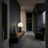 A modern, minimalist living space with dark hues; a floating wooden shelf displays books, a vase, and the Esmora 1 Light Marble Core Table Lamp in Grey & White. A curved armchair faces large floor-to-ceiling windows overlooking the city.